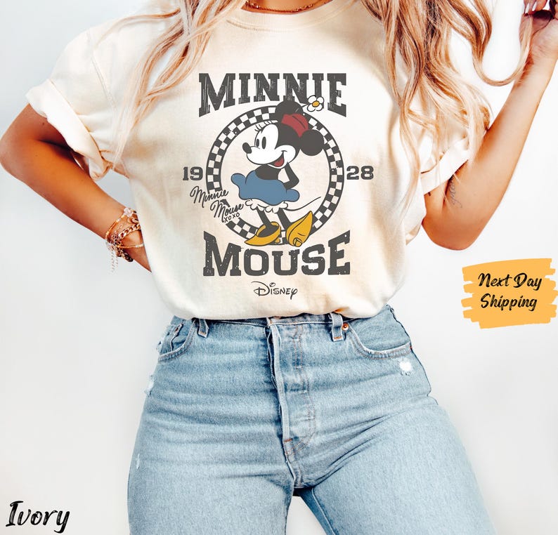 K&ouml;nnte beinhalten: Ein wei&szlig;es T-Shirt mit einem Vintage-Grafikdruck von Minnie Mouse. Die Grafik zeigt Minnie Mouse in einem roten Kleid und gelben Schuhen, mit dem Text "Minnie Mouse" und "Disney" darunter. Die Grafik ist von einem karierten Rand umgeben.