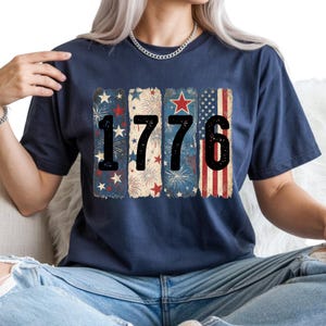 May include: Navy blue t-shirt with the number "1776" in black. The numbers are set against a backdrop of vertical stripes featuring stars, fireworks, and the American flag. The shirt is a casual wear item.