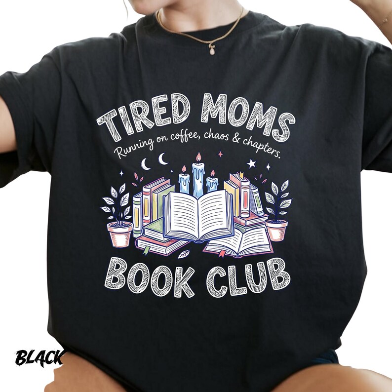 May include: Black t-shirt with the text "TIRED MOMS BOOK CLUB" in white. The design includes an illustration of books, candles, and plants. The phrase "Running on coffee, chaos & chapters" is also included.