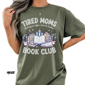May include: Moss green t-shirt with the text "TIRED MOMS BOOK CLUB" in a white, distressed font. The graphic features books, candles, and plants. The phrase "Running on coffee, chaos & chapters" is also included.