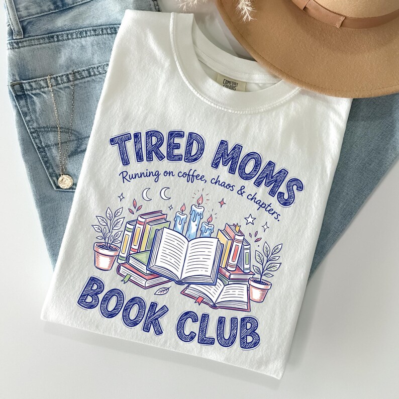 May include: White t-shirt with the text "TIRED MOMS BOOK CLUB" in blue. The shirt features a graphic of books, candles, and plants. The shirt is laid on top of blue jeans and a tan hat.