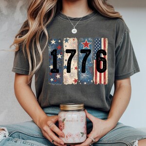 May include: Dark gray t-shirt with the number "1776" in black, with a patriotic design. The design includes red, white, and blue elements such as stars, fireworks, and stripes. The shirt is a comfortable, casual style.