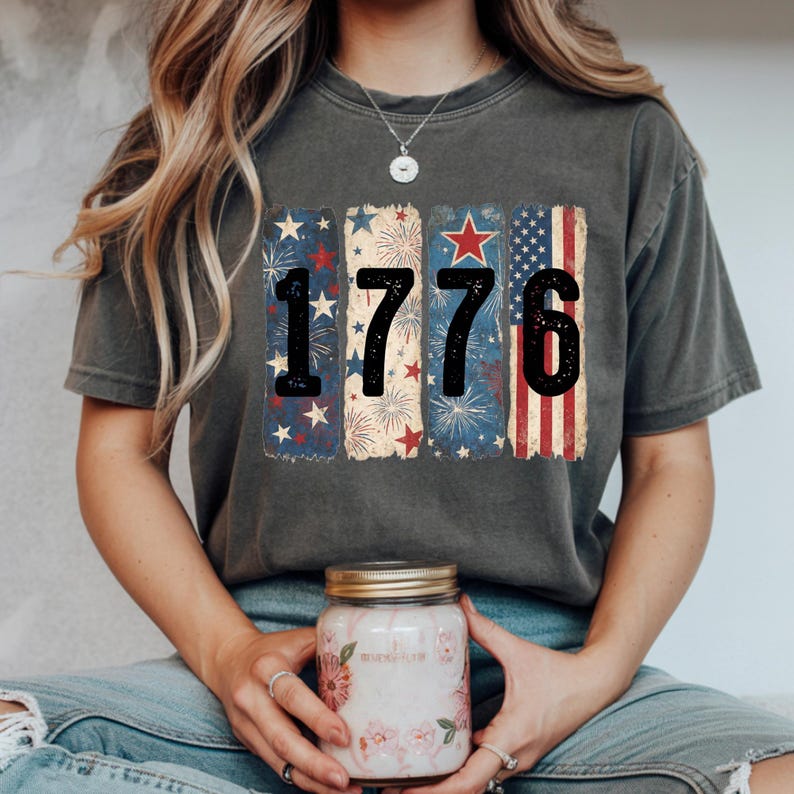 May include: Dark gray t-shirt with the number "1776" in black, with a patriotic design. The design includes red, white, and blue elements such as stars, fireworks, and stripes. The shirt is a comfortable, casual style.