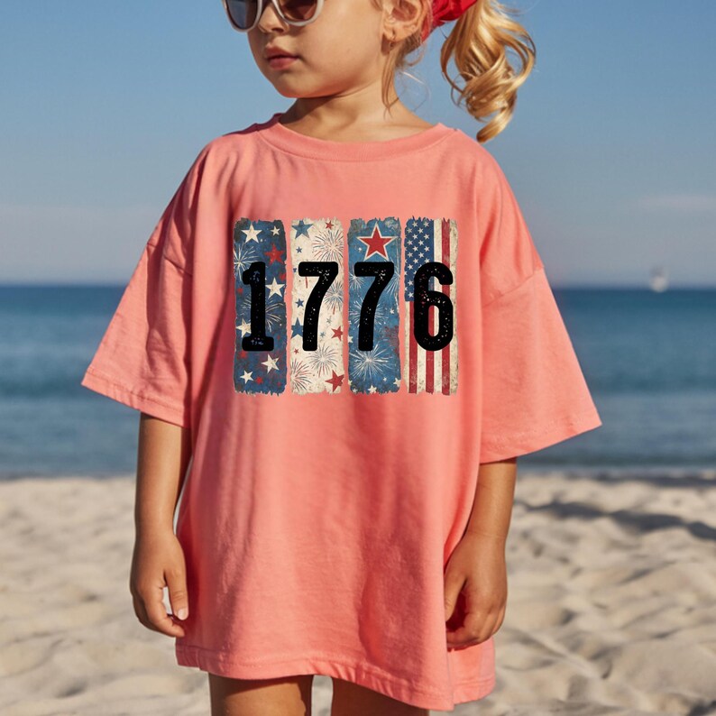 May include: A coral-colored t-shirt featuring the number "1776" in bold black font. The numbers are superimposed over vertical rectangular designs with patriotic motifs, including stars, fireworks, and stripes in red, white, and blue.