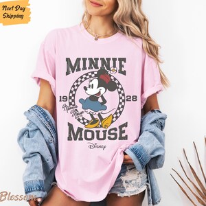 K&ouml;nnte beinhalten: Pinkes T-Shirt mit einem Vintage-Grafikdruck von Minnie Mouse. Die Grafik zeigt Minnie Mouse in einem schwarz-wei&szlig; karierten Kreis mit dem Text "Minnie Mouse" und "Disney" in einer verwitterten Schriftart.