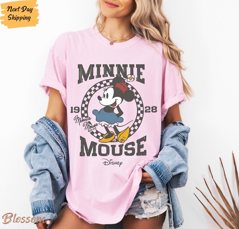 K&ouml;nnte beinhalten: Pinkes T-Shirt mit einem Vintage-Grafikdruck von Minnie Mouse. Die Grafik zeigt Minnie Mouse in einem schwarz-wei&szlig; karierten Kreis mit dem Text "Minnie Mouse" und "Disney" in einer verwitterten Schriftart.