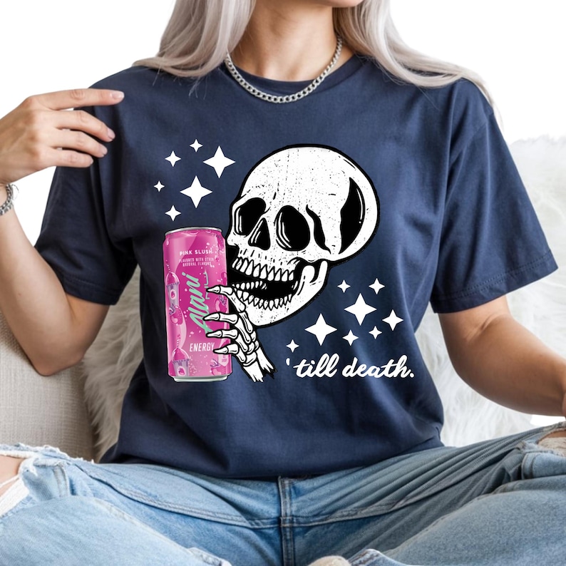 May include: Navy blue t-shirt featuring a white skull graphic holding a pink energy drink can. The design includes white stars and the phrase "'till death." The can has the text "Alani" and "Pink Slush" on it.