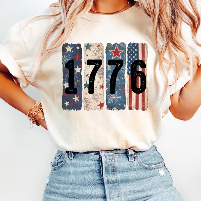 May include: Cream-colored t-shirt with the year "1776" printed in black. The numbers are set against vertical rectangular backgrounds featuring patriotic designs of stars, fireworks, and stripes in red, white, and blue.