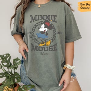 K&ouml;nnte beinhalten: Ein gr&uuml;nes T-Shirt mit einem Vintage-Grafikdruck von Minnie Mouse. Die Grafik zeigt Minnie Mouse in ihrem charakteristischen roten Kleid mit wei&szlig;en Punkten und dem Text "Minnie Mouse" und "1928" in einer verwitterten Schrift.