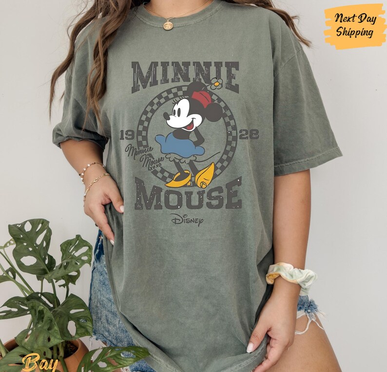 K&ouml;nnte beinhalten: Ein gr&uuml;nes T-Shirt mit einem Vintage-Grafikdruck von Minnie Mouse. Die Grafik zeigt Minnie Mouse in ihrem charakteristischen roten Kleid mit wei&szlig;en Punkten und dem Text "Minnie Mouse" und "1928" in einer verwitterten Schrift.