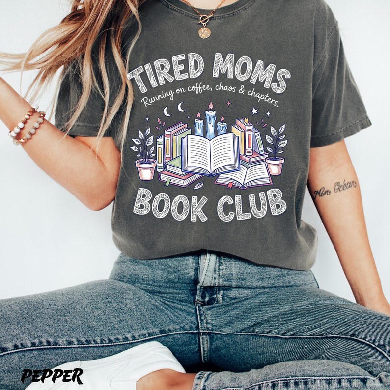 May include: Dark gray t-shirt with the text "TIRED MOMS BOOK CLUB" in white. The design includes illustrations of books, candles, and plants. The phrase "Running on coffee, chaos & chapters" is also printed on the shirt.