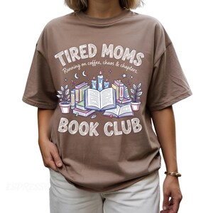 May include: Brown t-shirt with the text "TIRED MOMS BOOK CLUB" in a whimsical font. The design includes illustrations of books, candles, and plants. The shirt also has the phrase "Running on coffee, chaos & chapters."