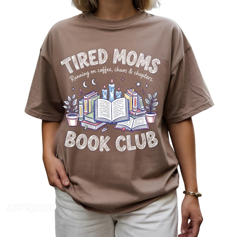 May include: Brown t-shirt with the text "TIRED MOMS BOOK CLUB" in a whimsical font. The design includes illustrations of books, candles, and plants. The shirt also has the phrase "Running on coffee, chaos & chapters."
