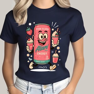 May include: Navy blue t-shirt featuring a cartoon illustration of a pink energy drink can with a smiling face, holding strawberry smoothies. The can is labeled "Alari Energy" and "Pink Slush". The shirt is a crew neck.