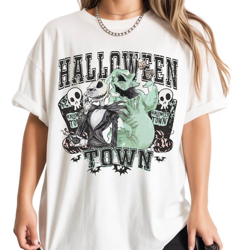 May include: A white t-shirt featuring a graphic print with the text HALLOWEEN TOWN. The design includes a skeletal figure in a black suit, a light green sack-like character, two skulls on tombstones, small pumpkins, and bats in black and green.