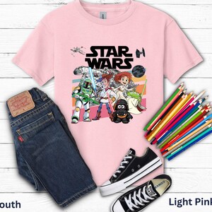 May include: Light pink youth t-shirt with a Star Wars graphic featuring characters and the Star Wars logo. The image includes a pair of blue jeans, black sneakers, and a set of colored pencils.