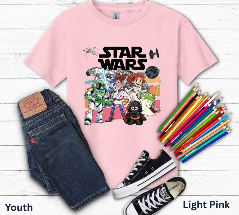 May include: Light pink youth t-shirt with a Star Wars graphic featuring characters and the Star Wars logo. The image includes a pair of blue jeans, black sneakers, and a set of colored pencils.
