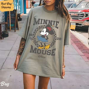 K&ouml;nnte beinhalten: Ein gr&uuml;nes Oversize-T-Shirt mit einem Vintage-Grafikdruck von Minnie Mouse. Die Grafik zeigt Minnie Mouse in einem blauen Kleid und gelben Schuhen, mit dem Text "Minnie Mouse" und "Disney" darunter.
