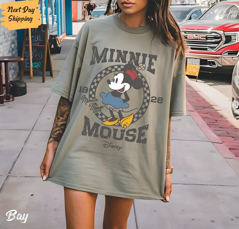 K&ouml;nnte beinhalten: Ein gr&uuml;nes Oversize-T-Shirt mit einem Vintage-Grafikdruck von Minnie Mouse. Die Grafik zeigt Minnie Mouse in einem blauen Kleid und gelben Schuhen, mit dem Text "Minnie Mouse" und "Disney" darunter.
