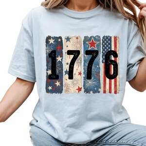 May include: Light blue t-shirt featuring the number "1776" in black, with each digit set against a patriotic design. The designs include stars, fireworks, and the American flag, creating a vintage, distressed look.