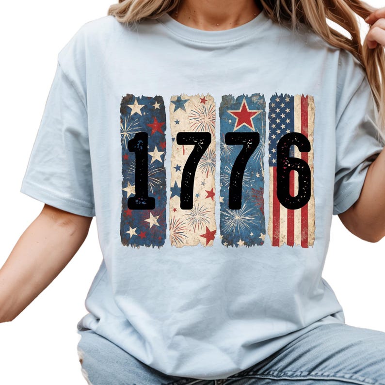 May include: Light blue t-shirt featuring the number "1776" in black, with each digit set against a patriotic design. The designs include stars, fireworks, and the American flag, creating a vintage, distressed look.