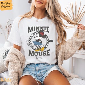 K&ouml;nnte beinhalten: Wei&szlig;es T-Shirt mit einem Vintage-Grafikdruck von Minnie Mouse. Die Grafik zeigt Minnie Mouse in ihrem charakteristischen gepunkteten Kleid und Schleife, mit dem Text "Minnie Mouse" und "1928" um sie herum. Der Text "Disney" ist ebenfalls in der Grafik enthalten.