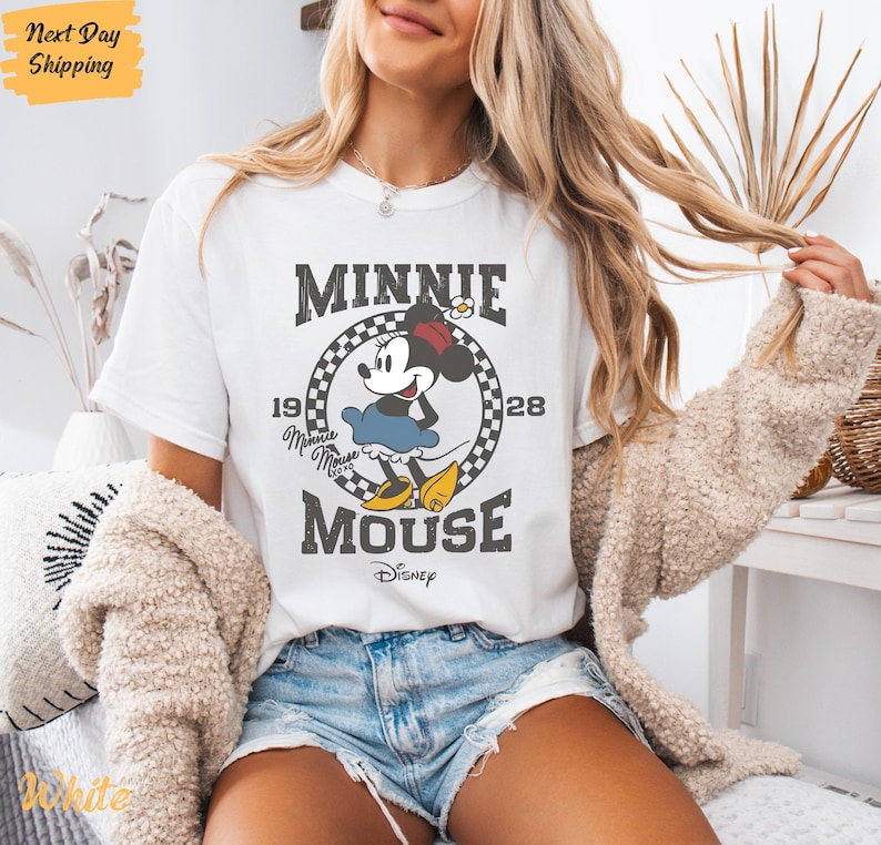 K&ouml;nnte beinhalten: Wei&szlig;es T-Shirt mit einem Vintage-Grafikdruck von Minnie Mouse. Die Grafik zeigt Minnie Mouse in ihrem charakteristischen gepunkteten Kleid und Schleife, mit dem Text "Minnie Mouse" und "1928" um sie herum. Der Text "Disney" ist ebenfalls in der Grafik enthalten.