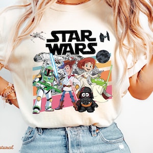 May include: Cream-colored Star Wars graphic t-shirt featuring characters from the Star Wars and Toy Story franchises. The design includes the Star Wars logo, a spaceship, and characters like Woody, Buzz Lightyear, and Darth Vader.