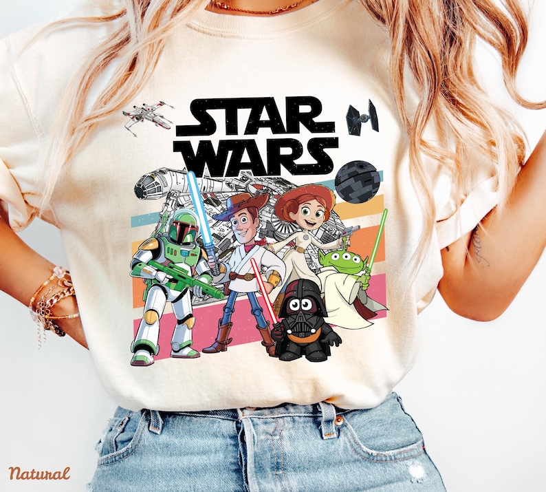 May include: Cream-colored Star Wars graphic t-shirt featuring characters from the Star Wars and Toy Story franchises. The design includes the Star Wars logo, a spaceship, and characters like Woody, Buzz Lightyear, and Darth Vader.