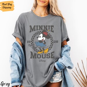 K&ouml;nnte beinhalten: Graues T-Shirt mit einem Vintage-Grafikdruck von Minnie Mouse. Die Grafik zeigt Minnie Mouse in einem blauen Kleid und roten Schuhen, mit dem Text "Minnie Mouse" und "Disney" darunter. Die Grafik ist von einem karierten Rahmen umgeben.