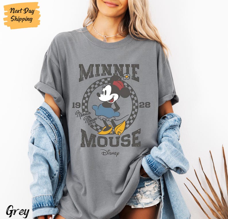 K&ouml;nnte beinhalten: Graues T-Shirt mit einem Vintage-Grafikdruck von Minnie Mouse. Die Grafik zeigt Minnie Mouse in einem blauen Kleid und roten Schuhen, mit dem Text "Minnie Mouse" und "Disney" darunter. Die Grafik ist von einem karierten Rahmen umgeben.