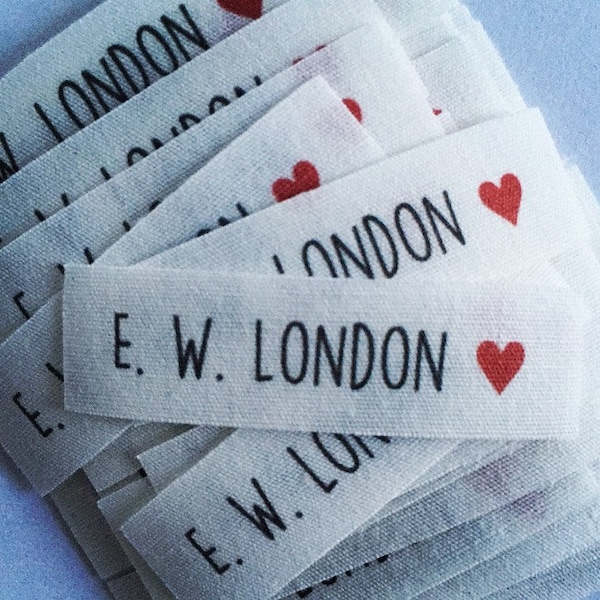 May include: Close-up of a pile of white fabric labels. Each label has the text "E. W. LONDON" in black, with a small red heart. The word "LONDON" is also printed on the labels. These labels are likely for clothing or craft projects.