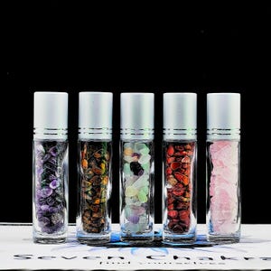 May include: Five clear glass roller bottles with silver caps filled with various colored crystals. The bottles are arranged in a row on a black surface.