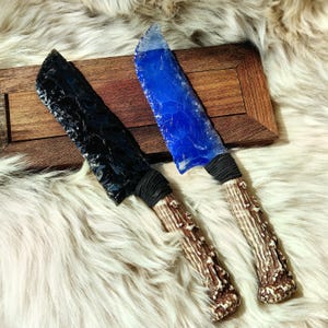 Puede incluir: Dos cuchillos con mangos de asta de ciervo. Un cuchillo tiene una hoja de obsidiana negra y el otro tiene una hoja de vidrio azul.