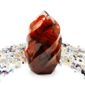 May include: A polished, flame-shaped carnelian crystal. The gemstone is a deep reddish-orange colour with swirling patterns and a glossy finish. The crystal is displayed on a white surface with a scattering of small, colourful crystals around its base.