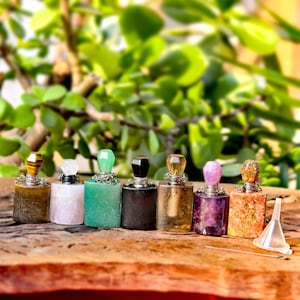May include: A collection of seven small, rectangular perfume bottles with gemstone stoppers. The bottles are arranged on a wooden surface, each with a unique color and stone, including tiger's eye, rose quartz, and amethyst. A small funnel is also present.