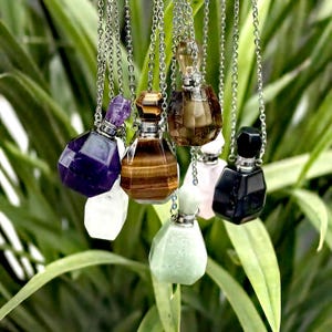 Może przedstawiać: Zestaw siedmiu butelek perfumowych z kryształów różnych kamieni szlachetnych na srebrnych łańcuszkach. Butelki są w różnych kolorach, w tym ametyst, tygrysie oko, przezroczysty, różowy kwarc i czarny onyks.