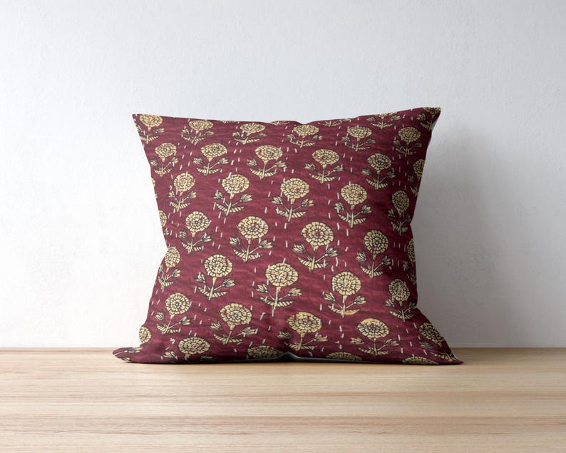 Puede incluir: Un coj&iacute;n cuadrado decorativo con un fondo burdeos y un patr&oacute;n floral repetido en color amarillo claro. Las flores tienen un dise&ntilde;o detallado, y el coj&iacute;n est&aacute; colocado sobre una superficie de madera.
