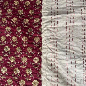 Puede incluir: Textil con dos patrones distintos. Un lado presenta un fondo rojo oscuro con un dise&ntilde;o floral dorado repetido. El otro lado es blanquecino con filas verticales de costuras rojas. Probablemente sea un edred&oacute;n o una manta.