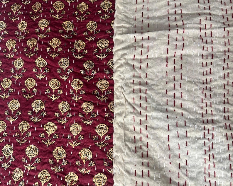 Puede incluir: Textil con dos patrones distintos. Un lado presenta un fondo rojo oscuro con un dise&ntilde;o floral dorado repetido. El otro lado es blanquecino con filas verticales de costuras rojas. Probablemente sea un edred&oacute;n o una manta.
