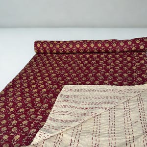 Puede incluir: Un edred&oacute;n burdeos con un estampado floral dorado. El edred&oacute;n est&aacute; parcialmente enrollado, revelando un reverso crema con costuras verticales. El estampado floral cubre la superficie superior.