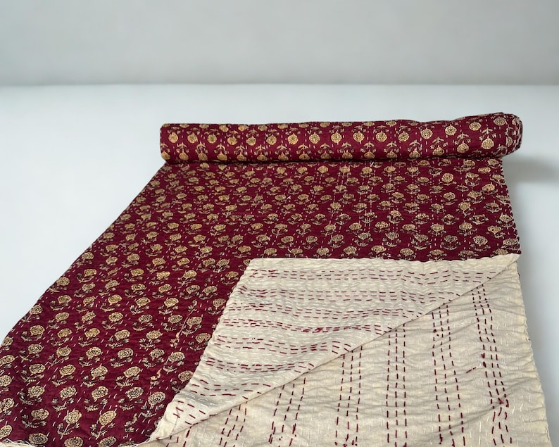 Puede incluir: Un edred&oacute;n burdeos con un estampado floral dorado. El edred&oacute;n est&aacute; parcialmente enrollado, revelando un reverso crema con costuras verticales. El estampado floral cubre la superficie superior.
