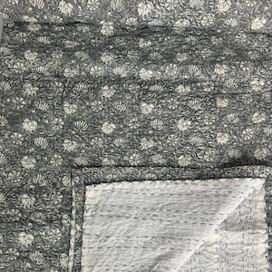 Peut inclure: Un textile matelassé rectangulaire présentant un fond gris foncé avec un motif répétitif de fleurs et de feuilles blanches. Le tissu affiche des détails de points de couture visibles sur toute la surface, avec un coin replié révélant un envers gris clair.