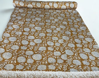 May include: A rectangular textile featuring a mustard yellow background with a repeating white floral pattern of large chrysanthemum-like blooms and leafy vines. The fabric has visible fine stitching throughout and is rolled at the top edge.