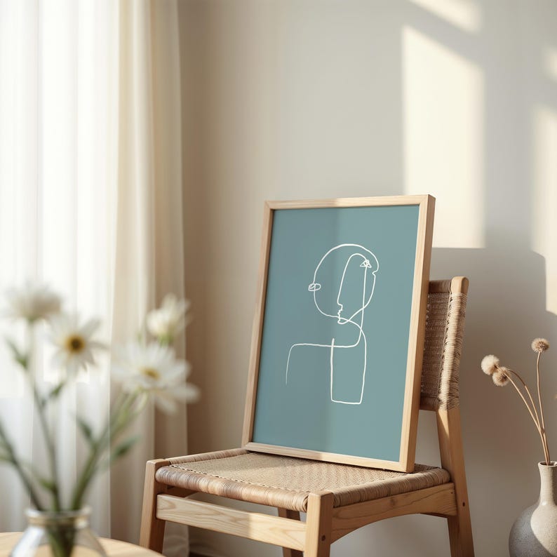 May include: A framed abstract line art print of a face in white on a teal background. The print is sitting on a wooden chair with a woven seat.