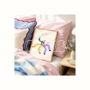 Op de afbeelding: Een ingelijste kunstprint met twee dansende figuren in groene, gele en paarse kleding. Het woord "golo" staat in roze geschreven bovenaan. De print ligt op een bed met roze en blauwe kussens en een patroon deken.