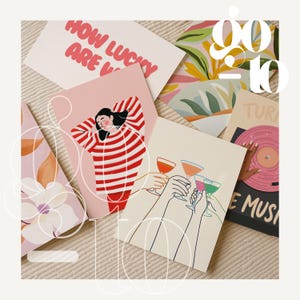 May include: A collection of greeting cards with various illustrations and text. One card reads "How Lucky Are V" in pink letters. Other cards feature images of people, cocktails, and a record player. The cards are arranged on a textured surface.