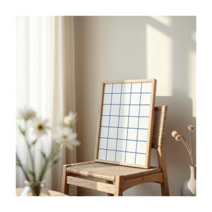 May include: A framed grid print with blue lines on a white background, leaning against a wooden chair with a woven seat. The chair is in a room with a white curtain and a vase with dried flowers.