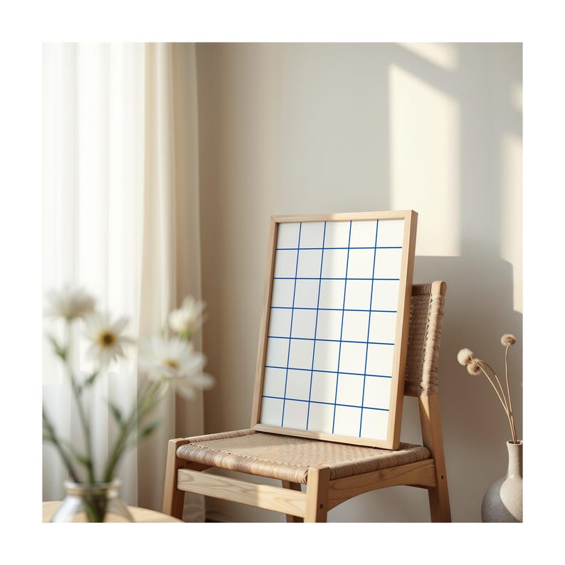 May include: A framed grid print with blue lines on a white background, leaning against a wooden chair with a woven seat. The chair is in a room with a white curtain and a vase with dried flowers.