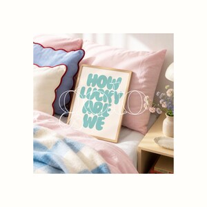 Op de afbeelding: Een ingelijste kunstprint met de tekst "HOW LUCKY ARE WE" in lichtblauw. De print ligt op een bed met roze kussens en een blauw-witte deken. Een kleine vaas met bloemen staat op een houten nachtkastje.